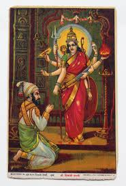 Devi stands wearing a red sari while a man kneels before her, appearing to pray.